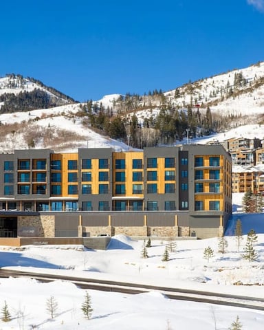 Large wooden lodge with multiple decks and stone foundation, surrounded by aspen trees and green lawn, with wooden bridge crossing a stream in foreground.